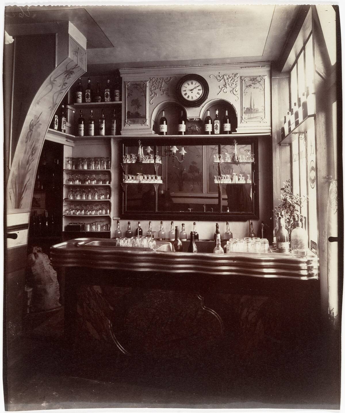 Photographie ancienne en noir et blanc de l'intérieur d'un marchand de vin, rue Boyer à Paris, vers 1910 : un comptoir en métal chargé de verres et de bouteilles, devant des étagères de verres, un grand miroir et une horloge.
