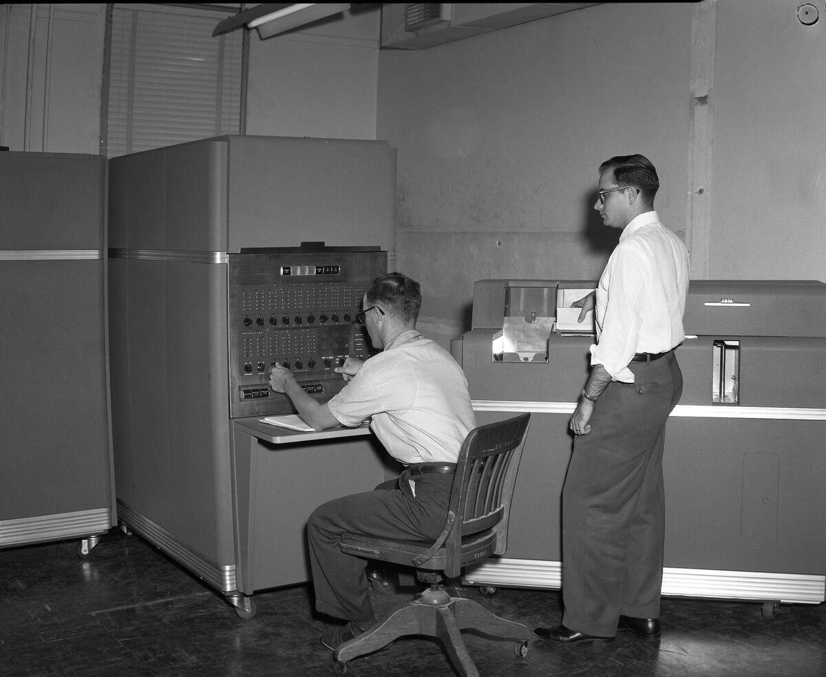 Photographie en noir et blanc : un homme assis règle les boutons du pupitre d'un grand ordinateur IBM 650, tandis qu'un second, debout, se tient près d'une autre machine marquée IBM.